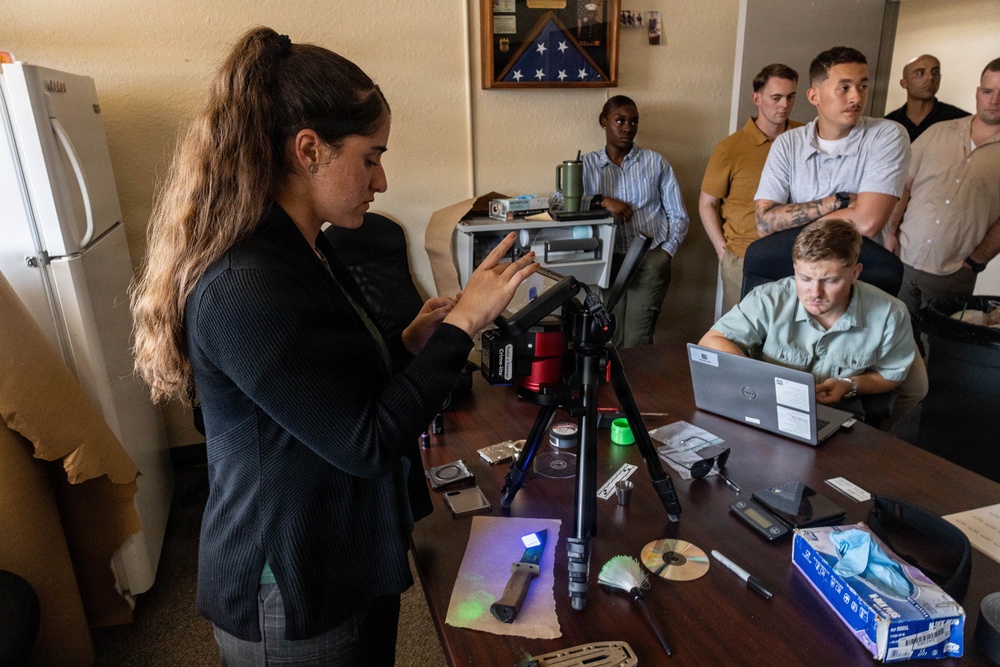 A woman is conducting what appears to be a forensic or investigative training demonstration for a group of observers in an office setting. She is operating a camera mounted on a tripod, with various forensic tools and evidence items spread across a desk. A group of approximately six individuals, likely trainees or students, are watching attentively.