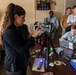 A woman is conducting what appears to be a forensic or investigative training demonstration for a group of observers in an office setting. She is operating a camera mounted on a tripod, with various forensic tools and evidence items spread across a desk. A group of approximately six individuals, likely trainees or students, are watching attentively.