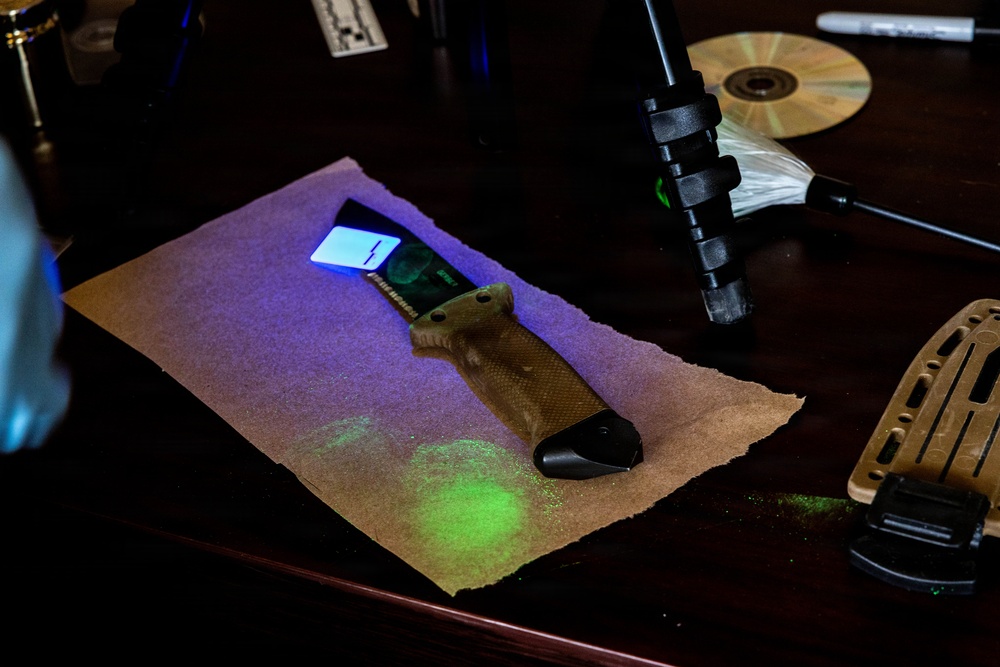 A forensic examination scene showing a tactical folding knife placed on brown paper under ultraviolet (UV) light illumination. Bright green fluorescent marks are visible on the paper beneath the knife, consistent with fingerprint powder or biological trace evidence reacting to UV light. The setting appears to be a forensic analysis workstation with various examination tools visible.