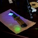 A forensic examination scene showing a tactical folding knife placed on brown paper under ultraviolet (UV) light illumination. Bright green fluorescent marks are visible on the paper beneath the knife, consistent with fingerprint powder or biological trace evidence reacting to UV light. The setting appears to be a forensic analysis workstation with various examination tools visible.