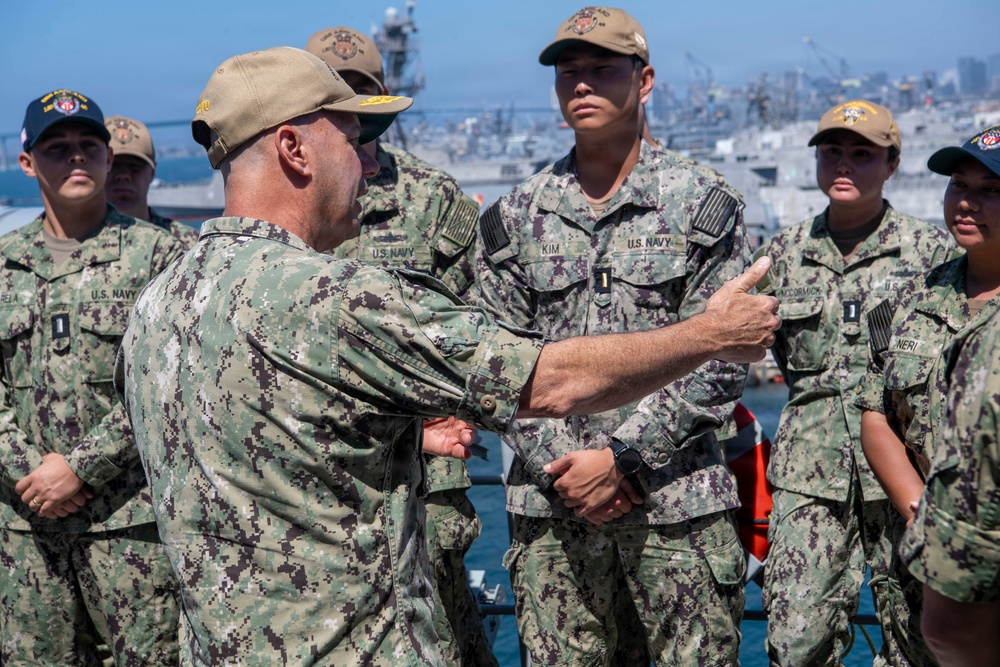 A senior U.S. Navy officer is addressing or inspecting a group of junior sailors/officers on what appears to be a naval pier or ship deck. The officer facing away from the camera appears to be gesturing or pointing at a sailor whose name tape reads 'KIM,' possibly conducting an inspection or briefing. The setting appears to be a naval base with ships and port infrastructure visible in the background.