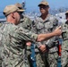 A senior U.S. Navy officer is addressing or inspecting a group of junior sailors/officers on what appears to be a naval pier or ship deck. The officer facing away from the camera appears to be gesturing or pointing at a sailor whose name tape reads 'KIM,' possibly conducting an inspection or briefing. The setting appears to be a naval base with ships and port infrastructure visible in the background.
