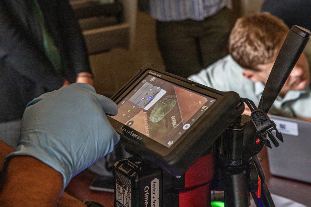 A gloved hand operates a Foster+Freeman forensic imaging device, specifically a Crime-lite AUTO unit mounted on a tripod. The tablet display shows what appears to be a forensic examination in progress, likely fingerprint or trace evidence analysis under specialized lighting. Several individuals are visible in the background observing the procedure.