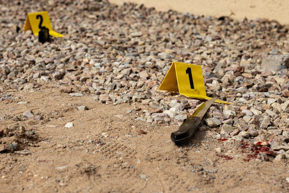 A crime scene investigation photograph showing an outdoor area with gravel and sandy ground. Two yellow forensic evidence markers (numbered 1 and 2) are visible, with marker 1 placed next to what appears to be a folding knife. A small area of reddish-brown staining consistent with blood is visible near marker 1.