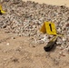 A crime scene investigation photograph showing an outdoor area with gravel and sandy ground. Two yellow forensic evidence markers (numbered 1 and 2) are visible, with marker 1 placed next to what appears to be a folding knife. A small area of reddish-brown staining consistent with blood is visible near marker 1.
