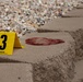 The image depicts a forensic crime scene evidence marker numbered '3', manufactured by Sirchie, placed adjacent to a dark reddish-brown stain on a concrete paver step. The stain appears irregular in shape and is consistent with a biological fluid deposit. The scene is set outdoors on a landscaped stairway bordered by decorative gravel.