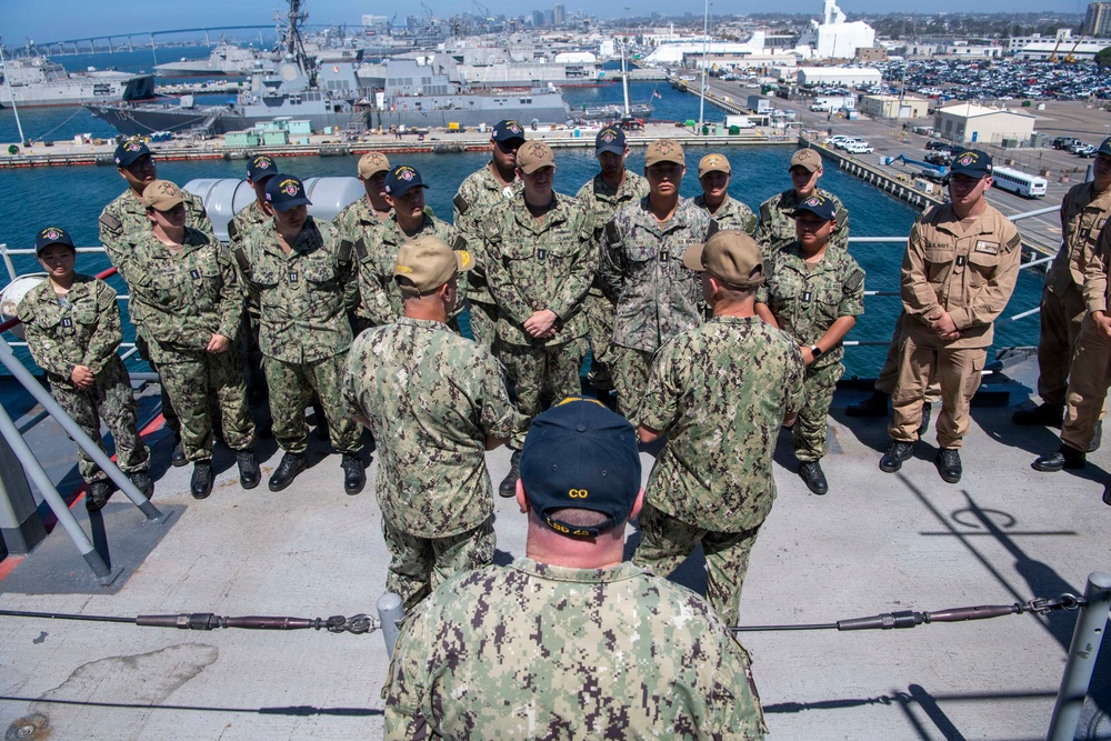 A group of U.S. Navy personnel are assembled on the deck of a naval vessel at what appears to be a military port or naval base. Two individuals with their backs to the camera, one wearing a cap labeled 'CO' (Commanding Officer) and another labeled 'LSD 25', appear to be addressing the assembled group. The background shows a busy naval installation with multiple ships, docks, and port infrastructure visible.
