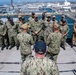 A group of U.S. Navy personnel are assembled on the deck of a naval vessel at what appears to be a military port or naval base. Two individuals with their backs to the camera, one wearing a cap labeled 'CO' (Commanding Officer) and another labeled 'LSD 25', appear to be addressing the assembled group. The background shows a busy naval installation with multiple ships, docks, and port infrastructure visible.