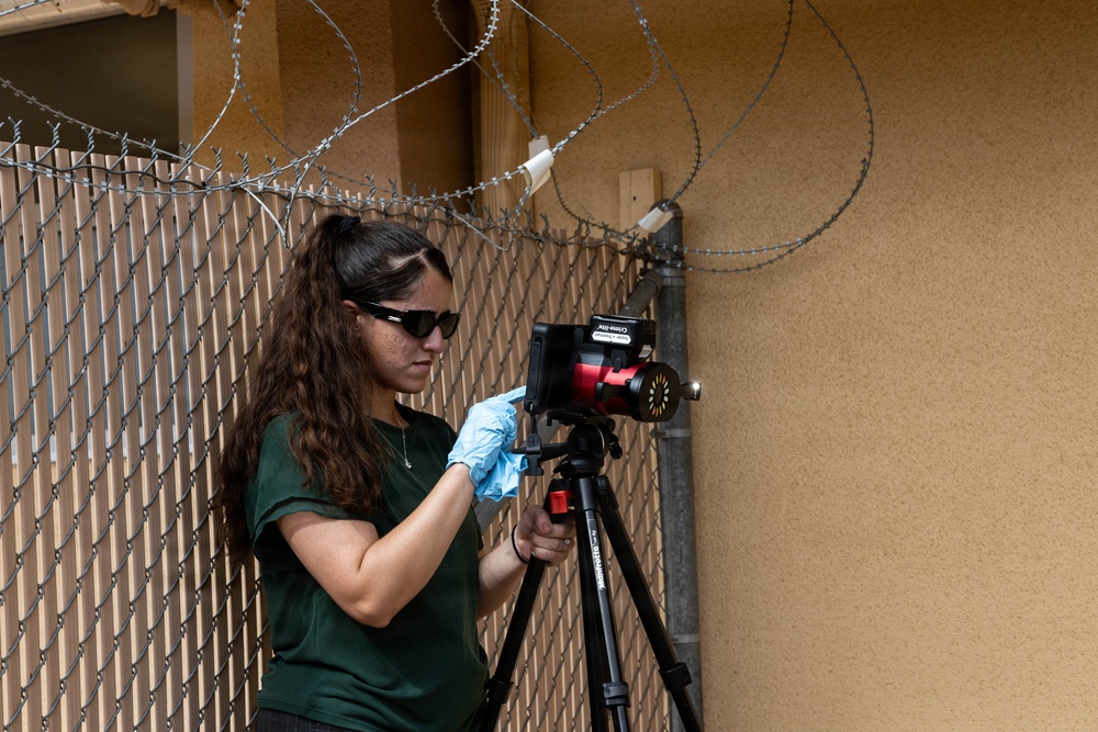 A young woman wearing sunglasses, a green t-shirt, and a blue latex glove is operating or adjusting a camera mounted on a black tripod. She is positioned near a chain-link fence topped with razor/concertina wire, with a tan stucco wall visible in the background. The setting suggests a secured or restricted perimeter area.