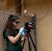 A young woman wearing sunglasses, a green t-shirt, and a blue latex glove is operating or adjusting a camera mounted on a black tripod. She is positioned near a chain-link fence topped with razor/concertina wire, with a tan stucco wall visible in the background. The setting suggests a secured or restricted perimeter area.
