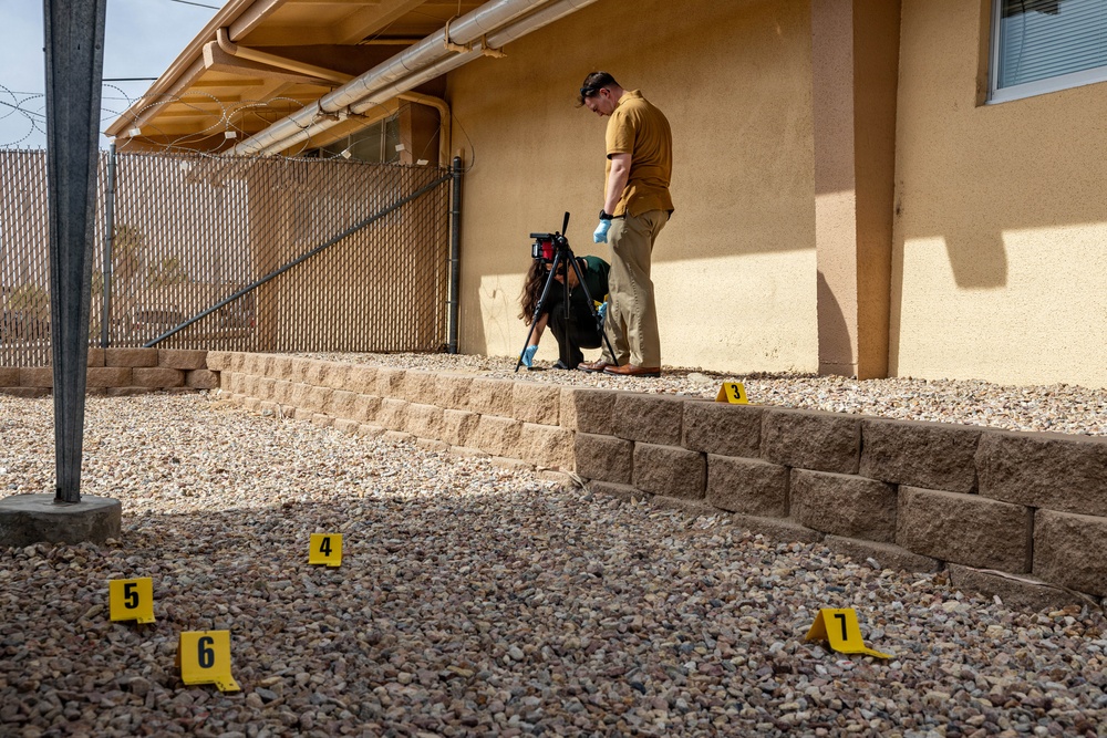 A crime scene investigation is underway at the exterior of a residential or commercial building. Two individuals wearing gloves are documenting the scene using a camera on a tripod, with multiple numbered yellow evidence markers (3, 4, 5, 6, 7) placed on a gravel-covered ground. The scene is located along the side of a stucco building near a chain-link fence topped with razor wire.