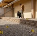A crime scene investigation is underway at the exterior of a residential or commercial building. Two individuals wearing gloves are documenting the scene using a camera on a tripod, with multiple numbered yellow evidence markers (3, 4, 5, 6, 7) placed on a gravel-covered ground. The scene is located along the side of a stucco building near a chain-link fence topped with razor wire.