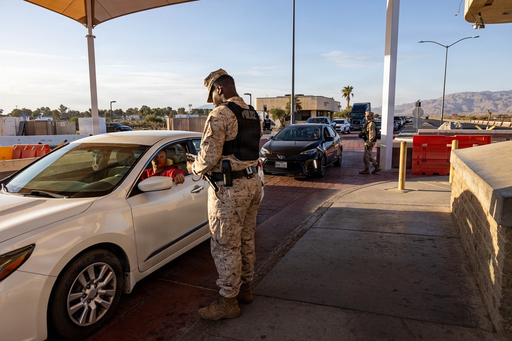 A military police officer in desert camouflage uniform and tactical vest is conducting a vehicle inspection at what appears to be a military base entry checkpoint. Multiple vehicles are queued in lanes under a covered checkpoint structure, with another uniformed personnel visible in the background. The setting includes desert terrain, palm trees, and mountains visible in the distance.