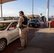 A military police officer in desert camouflage uniform and tactical vest is conducting a vehicle inspection at what appears to be a military base entry checkpoint. Multiple vehicles are queued in lanes under a covered checkpoint structure, with another uniformed personnel visible in the background. The setting includes desert terrain, palm trees, and mountains visible in the distance.
