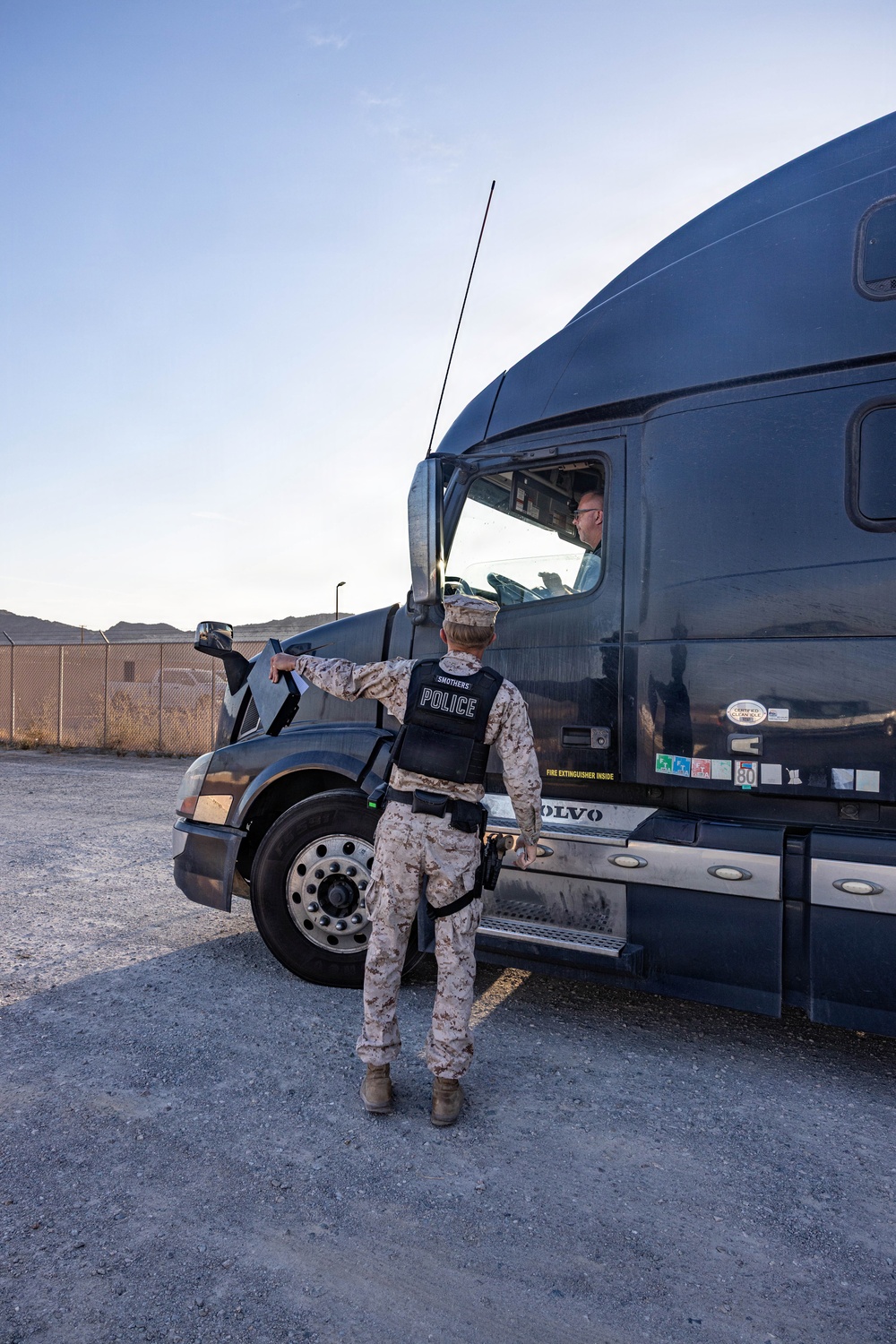 A military police officer in desert camouflage uniform and a black 'POLICE' vest is conducting a vehicle inspection or checkpoint stop of a dark-colored Volvo semi-truck. The officer, identified by the name 'SMOTHERS' on their vest, is standing beside the truck's cab and appears to be communicating with the driver. The scene appears to take place at a military installation gate or checkpoint, evidenced by the fencing and arid terrain in the background.