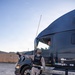 A military police officer in desert camouflage uniform and a black 'POLICE' vest is conducting a vehicle inspection or checkpoint stop of a dark-colored Volvo semi-truck. The officer, identified by the name 'SMOTHERS' on their vest, is standing beside the truck's cab and appears to be communicating with the driver. The scene appears to take place at a military installation gate or checkpoint, evidenced by the fencing and arid terrain in the background.