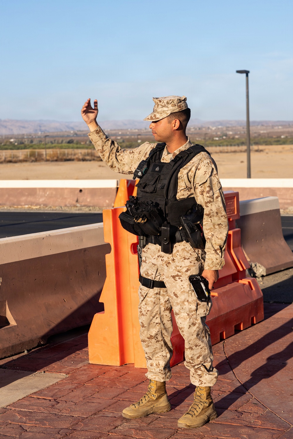 A U.S. Marine in desert MARPAT (MCCUU) uniform and tactical vest is standing at what appears to be a checkpoint or entry control point, raising one hand in a halt or signaling gesture. The setting includes orange traffic barriers, a concrete barrier wall, and an arid landscape in the background. The Marine is equipped with a sidearm, radio, and tactical gear consistent with a security or gate guard role.