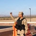 A U.S. Marine in desert MARPAT (MCCUU) uniform and tactical vest is standing at what appears to be a checkpoint or entry control point, raising one hand in a halt or signaling gesture. The setting includes orange traffic barriers, a concrete barrier wall, and an arid landscape in the background. The Marine is equipped with a sidearm, radio, and tactical gear consistent with a security or gate guard role.