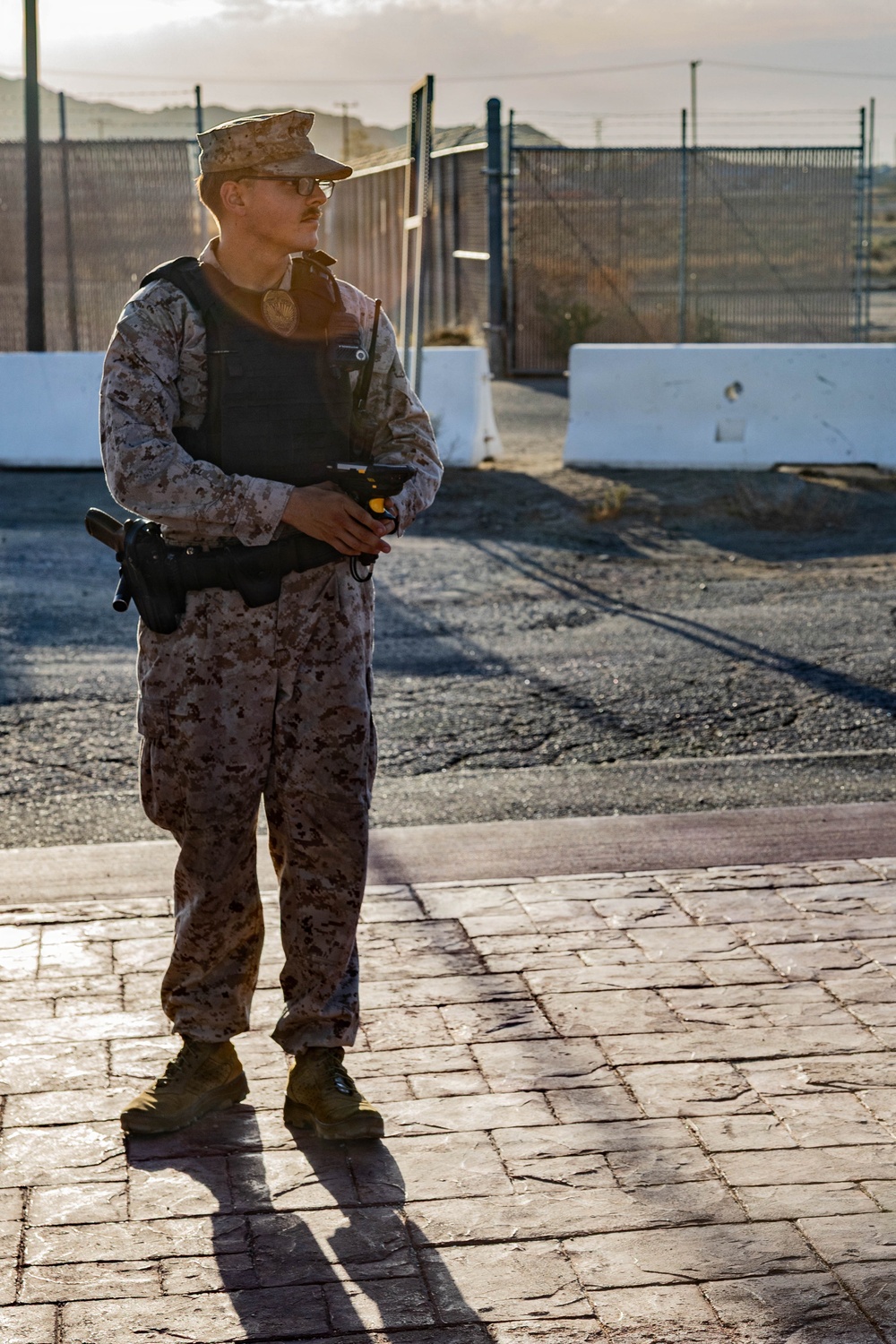 A military or security personnel member stands at what appears to be a base or facility entrance checkpoint in a desert environment. The individual is wearing desert camouflage (MARPAT pattern) uniform with a tactical vest, badge, and is holding a handheld device. The setting features concrete barriers, chain-link fencing, and arid terrain in the background.