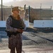 A military or security personnel member stands at what appears to be a base or facility entrance checkpoint in a desert environment. The individual is wearing desert camouflage (MARPAT pattern) uniform with a tactical vest, badge, and is holding a handheld device. The setting features concrete barriers, chain-link fencing, and arid terrain in the background.