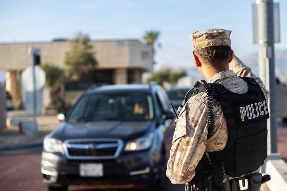 A military police officer in desert camouflage uniform and black tactical vest is manning what appears to be a security checkpoint or gate. The officer is facing away from the camera, monitoring a dark-colored Subaru SUV that has stopped in front of them. The setting appears to be a military installation entrance in an arid, desert environment.