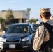 A military police officer in desert camouflage uniform and black tactical vest is manning what appears to be a security checkpoint or gate. The officer is facing away from the camera, monitoring a dark-colored Subaru SUV that has stopped in front of them. The setting appears to be a military installation entrance in an arid, desert environment.
