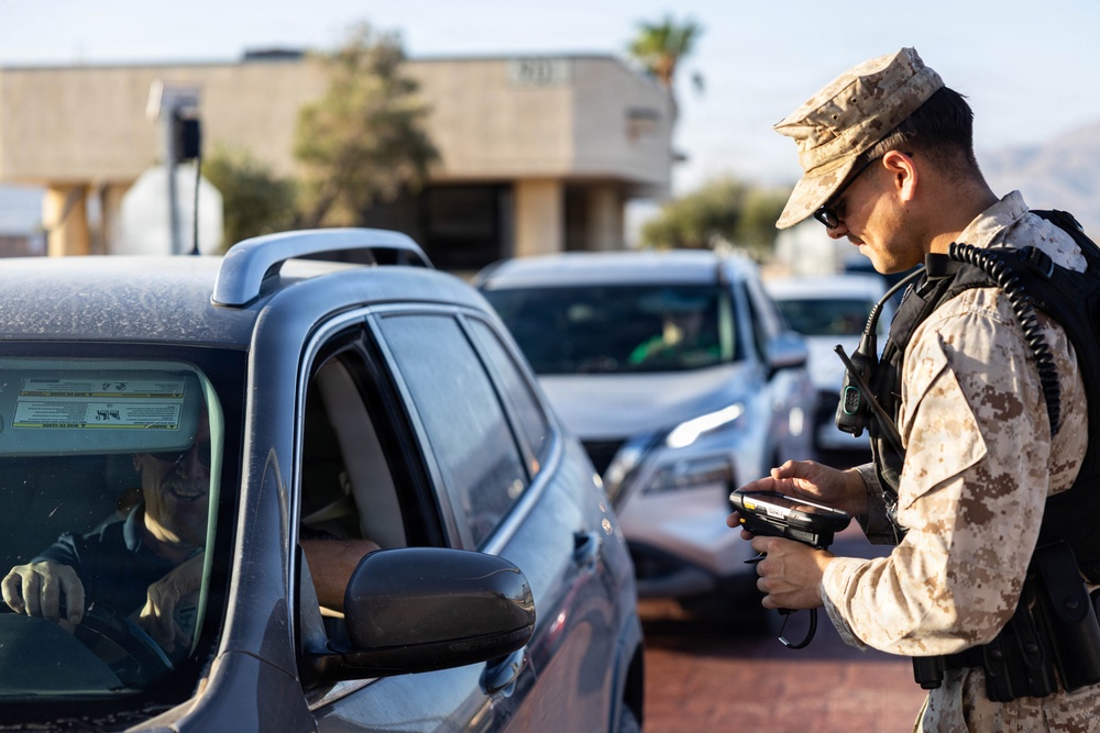 A military personnel wearing desert camouflage (MARPAT pattern) uniform is operating a handheld scanning device at what appears to be a vehicle checkpoint or base entry control point. The soldier is interacting with a vehicle driver while processing credentials or scanning documents. Multiple vehicles are queued in the background, suggesting an active access control operation.