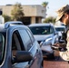 A military personnel wearing desert camouflage (MARPAT pattern) uniform is operating a handheld scanning device at what appears to be a vehicle checkpoint or base entry control point. The soldier is interacting with a vehicle driver while processing credentials or scanning documents. Multiple vehicles are queued in the background, suggesting an active access control operation.