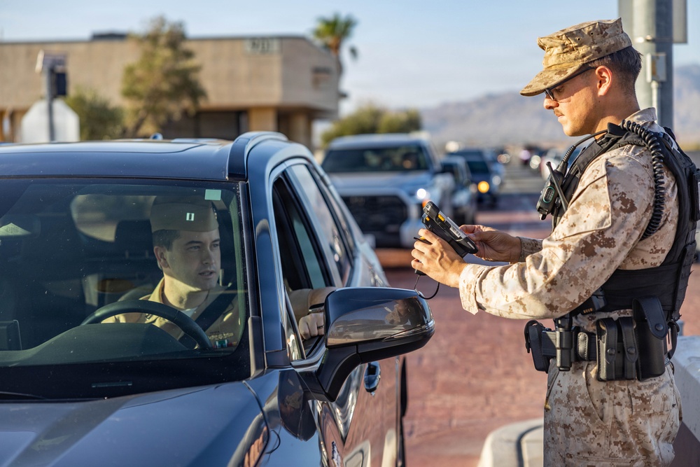 A uniformed military personnel wearing desert camouflage (MARPAT pattern) is conducting a vehicle checkpoint inspection, scanning or processing credentials with a handheld electronic device. The driver of a dark-colored vehicle is waiting while the guard reviews the information. A queue of vehicles is visible in the background, suggesting an active military base entry control point.