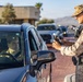 A uniformed military personnel wearing desert camouflage (MARPAT pattern) is conducting a vehicle checkpoint inspection, scanning or processing credentials with a handheld electronic device. The driver of a dark-colored vehicle is waiting while the guard reviews the information. A queue of vehicles is visible in the background, suggesting an active military base entry control point.