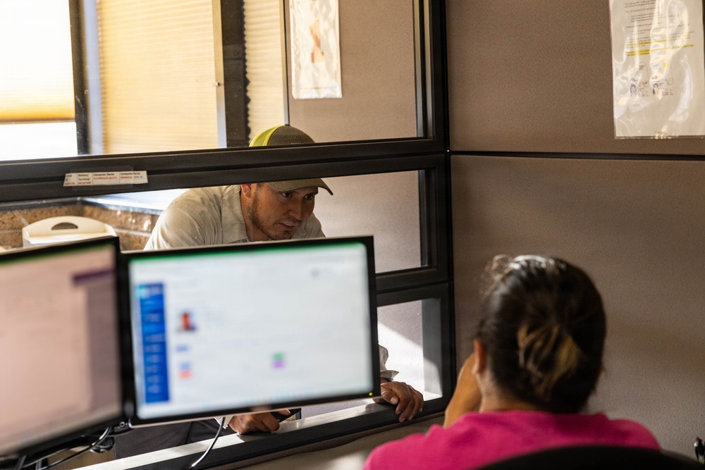 A man wearing a baseball cap and light-colored shirt is leaning toward a service window, speaking with a female clerk seated at a workstation. The clerk, wearing a pink top with her hair in a bun, faces two computer monitors displaying what appears to be a database or records management application. The setting appears to be a government office or administrative service counter, possibly a DMV, clinic, or similar public-facing agency.