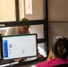 A man wearing a baseball cap and light-colored shirt is leaning toward a service window, speaking with a female clerk seated at a workstation. The clerk, wearing a pink top with her hair in a bun, faces two computer monitors displaying what appears to be a database or records management application. The setting appears to be a government office or administrative service counter, possibly a DMV, clinic, or similar public-facing agency.