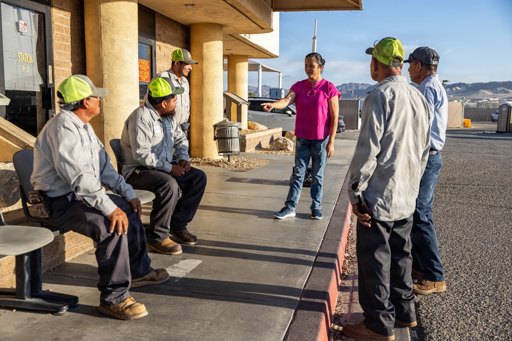 A woman in a pink shirt is addressing a group of five male workers wearing green caps and gray work uniforms outside a building labeled 'Station 1-3'. She appears to be giving instructions or conducting a briefing, pointing toward the workers. The setting appears to be an outdoor work facility or depot in a desert environment.