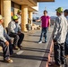 A woman in a pink shirt is addressing a group of five male workers wearing green caps and gray work uniforms outside a building labeled 'Station 1-3'. She appears to be giving instructions or conducting a briefing, pointing toward the workers. The setting appears to be an outdoor work facility or depot in a desert environment.