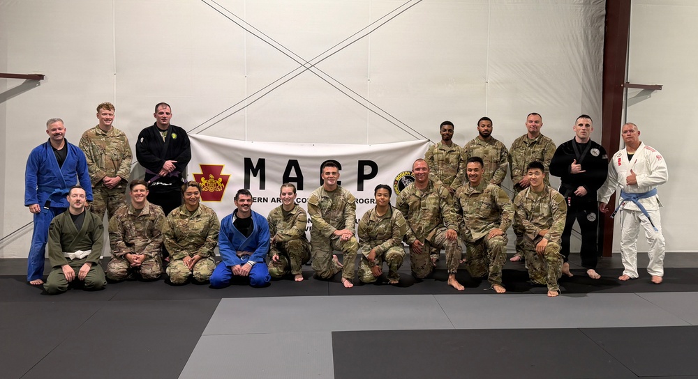 A group of approximately 20 individuals pose for a group photo in front of a banner reading 'MARP' (Modern Army Combatives Program) on a martial arts training mat. The group consists of military personnel in camouflage uniforms mixed with civilians wearing Brazilian Jiu-Jitsu gis in blue, white, and black. The setting appears to be an indoor martial arts training facility.