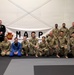 A group of approximately 20 individuals pose for a group photo in front of a banner reading 'MARP' (Modern Army Combatives Program) on a martial arts training mat. The group consists of military personnel in camouflage uniforms mixed with civilians wearing Brazilian Jiu-Jitsu gis in blue, white, and black. The setting appears to be an indoor martial arts training facility.