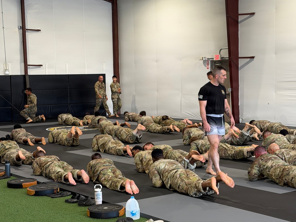 A large group of military personnel in camouflage uniforms are performing floor exercises on padded mats inside an indoor training facility. An instructor wearing a black t-shirt and shorts is walking among the trainees, overseeing the session. The setting appears to be a dedicated combatives or physical training gym with wall padding and exercise equipment visible.