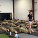 A large group of military personnel in camouflage uniforms are performing floor exercises on padded mats inside an indoor training facility. An instructor wearing a black t-shirt and shorts is walking among the trainees, overseeing the session. The setting appears to be a dedicated combatives or physical training gym with wall padding and exercise equipment visible.
