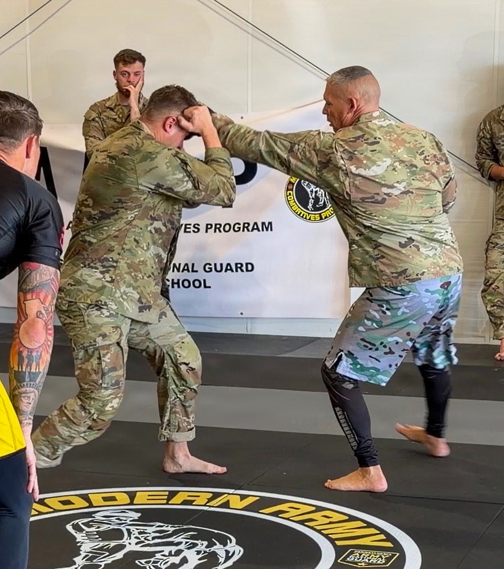 Two soldiers in U.S. Army OCP (Operational Camouflage Pattern) uniforms are engaged in a combatives training drill on a marked mat. One individual wearing mixed athletic/uniform attire is demonstrating a striking or clinch technique on a uniformed soldier. The setting is an official Army National Guard Combatives Program training facility.