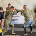 Two soldiers in U.S. Army OCP (Operational Camouflage Pattern) uniforms are engaged in a combatives training drill on a marked mat. One individual wearing mixed athletic/uniform attire is demonstrating a striking or clinch technique on a uniformed soldier. The setting is an official Army National Guard Combatives Program training facility.