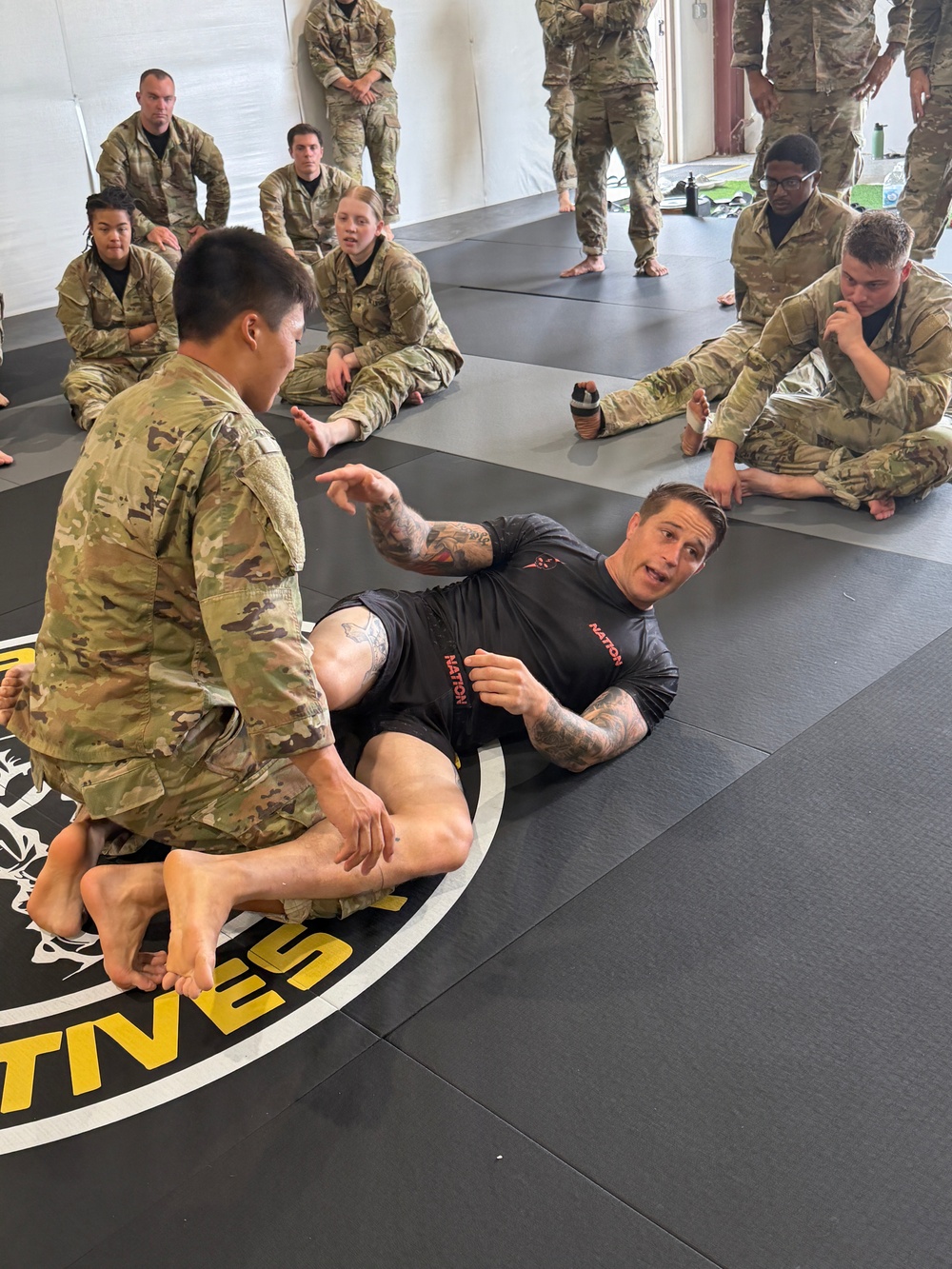 A martial arts or combatives instructor wearing a black 'NATION' branded rash guard is demonstrating a ground fighting technique to a group of military personnel in U.S. Army OCP (Operational Camouflage Pattern) uniforms. The instructor is on his back on a black mat, appearing to explain a guard or leg entanglement position to a soldier kneeling over him. Multiple soldiers are seated around the perimeter of the mat observing the demonstration.