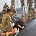 A martial arts or combatives instructor wearing a black 'NATION' branded rash guard is demonstrating a ground fighting technique to a group of military personnel in U.S. Army OCP (Operational Camouflage Pattern) uniforms. The instructor is on his back on a black mat, appearing to explain a guard or leg entanglement position to a soldier kneeling over him. Multiple soldiers are seated around the perimeter of the mat observing the demonstration.