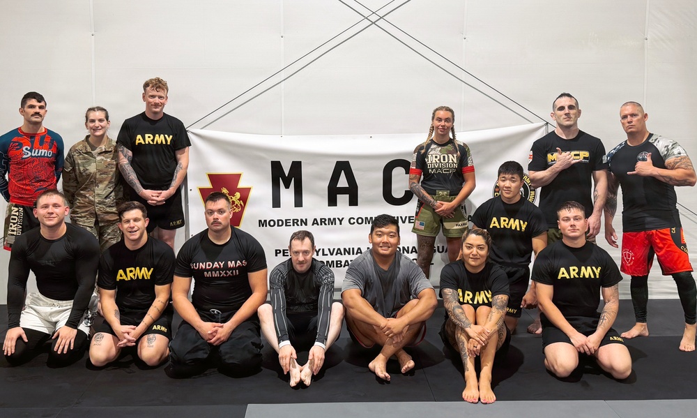 A group of approximately 15 individuals pose for a group photo in front of a banner reading 'MAC - Modern Army Combatives' with a Pennsylvania National Guard reference. The group includes military personnel in Army-branded athletic wear and civilian participants in grappling/MMA attire. The setting appears to be an indoor martial arts training facility with padded flooring.