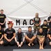 A group of approximately 15 individuals pose for a group photo in front of a banner reading 'MAC - Modern Army Combatives' with a Pennsylvania National Guard reference. The group includes military personnel in Army-branded athletic wear and civilian participants in grappling/MMA attire. The setting appears to be an indoor martial arts training facility with padded flooring.
