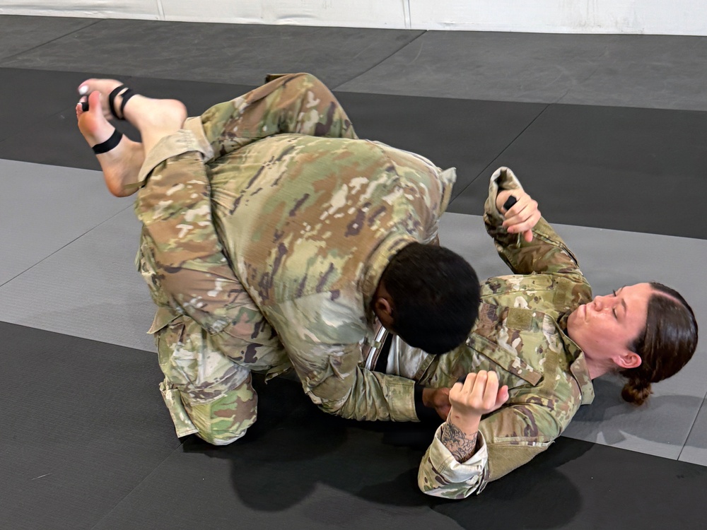 Two individuals wearing military camouflage uniforms (OCP/MultiCam pattern) are engaged in ground grappling on black martial arts mats. One person is on top in a dominant position while the other is on their back in a defensive posture. This appears to be a combatives training session, likely U.S. Army combatives or similar military hand-to-hand combat training.