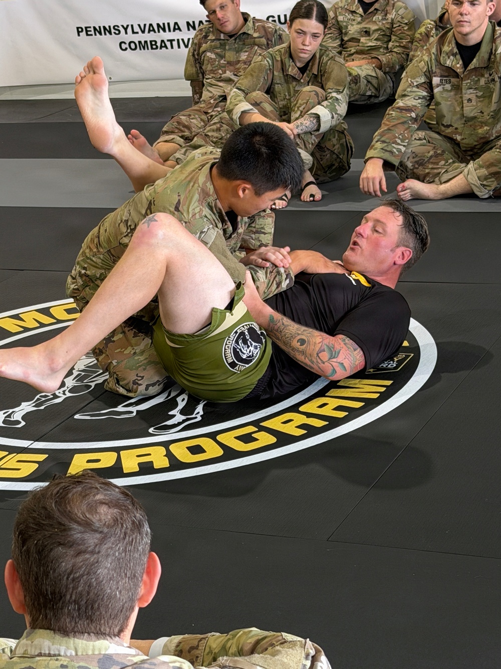 Two individuals are engaged in a ground combatives demonstration on a marked training mat, with one person in military camouflage uniform and the other in a black shirt applying or defending a grappling technique. They are surrounded by multiple soldiers in U.S. Army OCP (Operational Camouflage Pattern) uniforms who are seated and observing the demonstration. A banner in the background reads 'Pennsylvania National Guard Combatives Program.'