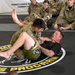Two individuals are engaged in a ground combatives demonstration on a marked training mat, with one person in military camouflage uniform and the other in a black shirt applying or defending a grappling technique. They are surrounded by multiple soldiers in U.S. Army OCP (Operational Camouflage Pattern) uniforms who are seated and observing the demonstration. A banner in the background reads 'Pennsylvania National Guard Combatives Program.'