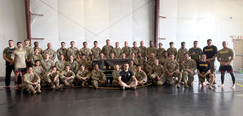 A large group of military personnel posed for a formal group photo inside what appears to be a training facility or gymnasium. The majority are wearing Army Combat Uniforms (ACU/OCP pattern), while several individuals on the edges wear black 'MACP' branded athletic shirts. They are gathered around a banner reading 'MACP - Modern Army Combatives - Pennsylvania [unit] Combatives.'