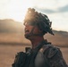 A military service member is photographed in a desert environment, appearing to shout or call out commands. The individual is wearing full tactical gear including a camouflage helmet with netting/ghillie material, tactical eyewear, and MARPAT (Marine pattern) camouflage uniform. The image is dramatically backlit by a setting or rising sun, creating a striking silhouette effect against an arid, mountainous landscape.