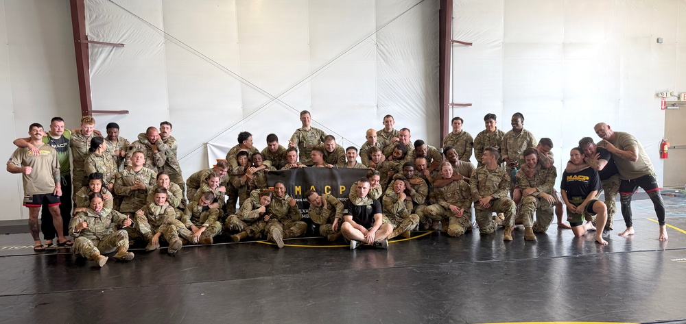 A large group of military personnel, predominantly in camouflage uniforms, pose for a group photo inside what appears to be a training facility or hangar. They are gathered around a black banner reading 'MACP' (Modern Army Combatives Program). The group includes both uniformed soldiers and a few individuals in civilian/athletic clothing, suggesting a combatives training event or course completion.
